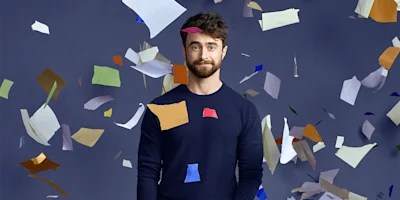 A man with a beard stands against a dark background as pieces of colorful paper float around him.