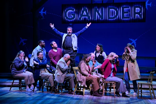 A group of actors sit on chairs on stage, reacting with surprise or concern, while one person stands with arms raised under a sign reading "GANDER" and airplane graphics in the background.