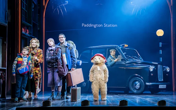 Actors on stage portray a family with luggage, standing next to a costumed Paddington Bear and a black taxi, with a “Paddington Station” sign in the background.