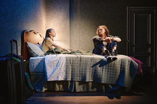 Two young women sit on a bed in a dimly lit room, engaged in conversation, with a suitcase nearby and soft lighting illuminating the scene.
