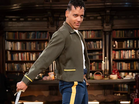 A person in a green jacket with yellow trim and blue pants holds a sword, standing in front of bookshelves in a library setting.