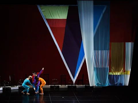 Four dancers in colorful bodysuits perform on stage in front of large geometric fabric panels and empty chairs against a dark backdrop.