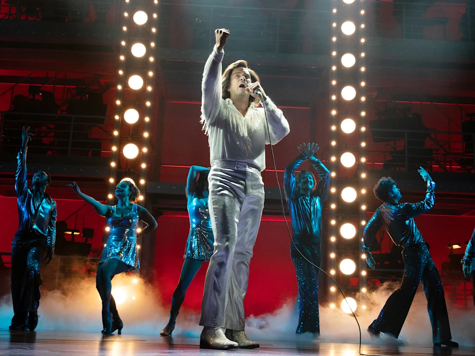 A singer in a white outfit performs on stage with backup dancers in blue costumes, illuminated by stage lights and surrounded by stage fog.