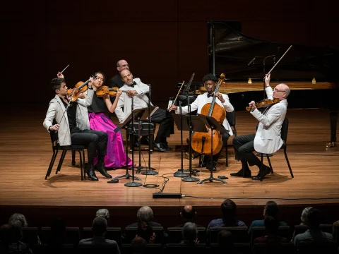 A string quintet performs on stage. Three violinists, one cellist, and a pianist, all dressed formally, play their instruments while seated. Audience members watch from the foreground.