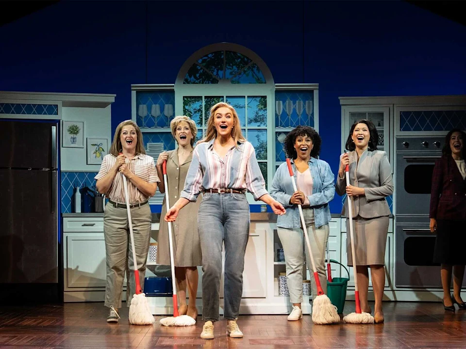 Five women stand on a stage set as a kitchen, four holding mops, while the woman in the center sings with a joyful expression.