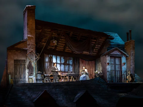 A stage set depicting an attic with actors in period costumes. One performer stands near a table, while the other sits with a sewing machine. The building's wooden roof is partially open.