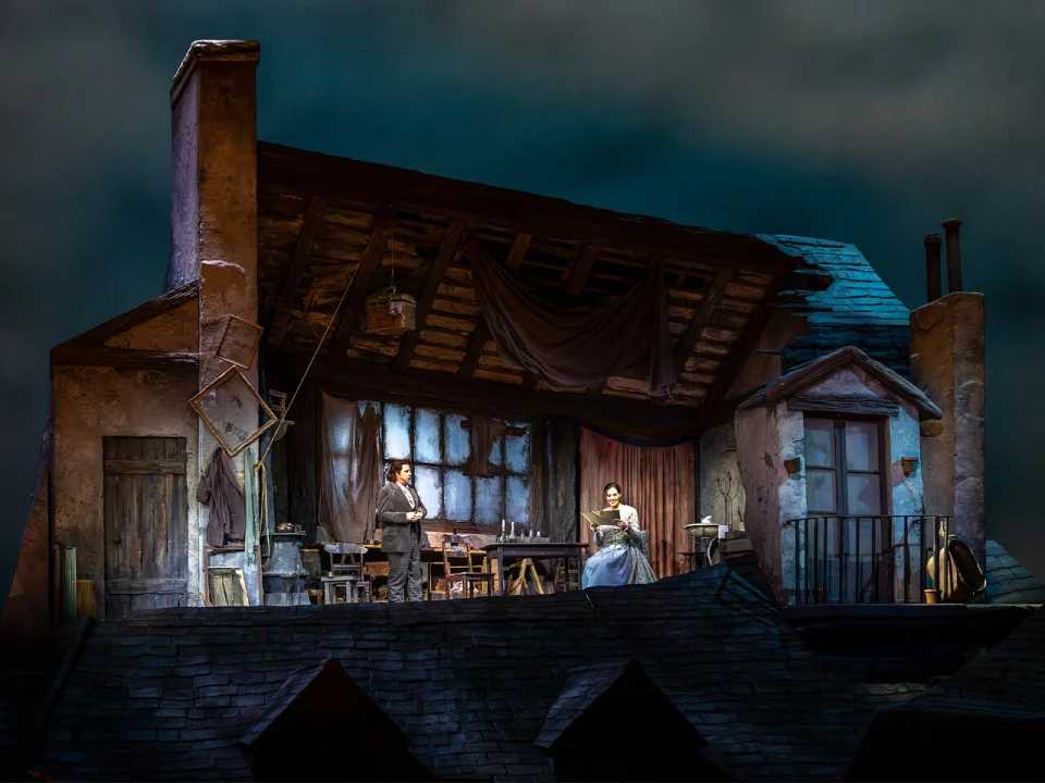 A stage set depicting an attic with actors in period costumes. One performer stands near a table, while the other sits with a sewing machine. The building's wooden roof is partially open.