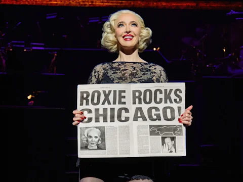 Person in stage costume holds a newspaper with the headline "Roxie Rocks Chicago" under stage lighting.