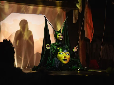 A performer in a black costume with large headpiece and a painted face on their chest sits on stage, with dramatic lighting and fabric backdrops.