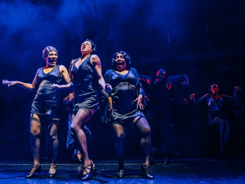 Performers in black costumes dance energetically on a dimly lit stage with blue lighting and fog effect surrounding them.