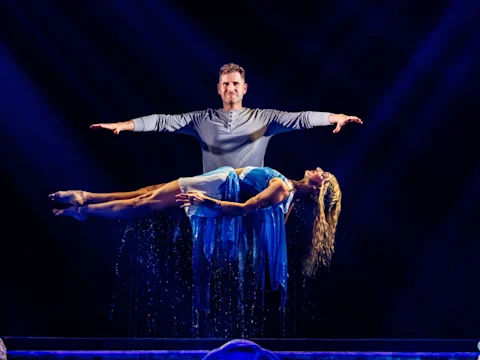 Production photo of Rob Lake Magic with Special Guests The Muppets on Broadway showing Rob Lake stands with arms outstretched as a woman in a blue dress appears to levitate horizontally in front of him on a dimly lit stage.