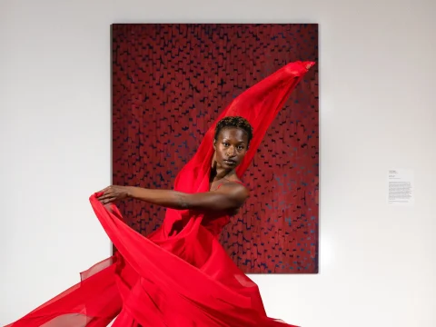 A person in a sweeping red dress performs a dance in front of a red and black abstract painting on a white wall.