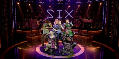Six performers in colorful, modern costumes pose on stage under purple lights, with a lit sign reading “SIX” and musicians playing instruments in the background.