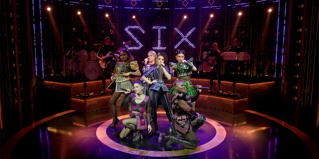 Six performers in colorful, modern costumes pose on stage under purple lights, with a lit sign reading “SIX” and musicians playing instruments in the background.