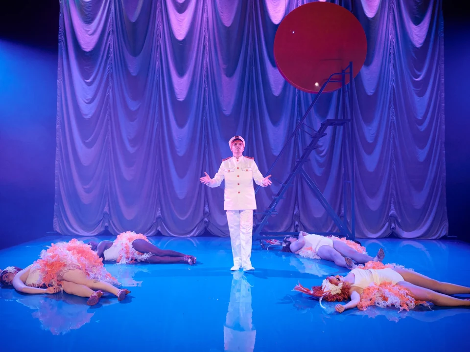 A person in a white naval uniform stands center stage, surrounded by four performers lying on the floor with pink feather boas. Behind them is a large red disc and a metallic curtain backdrop.