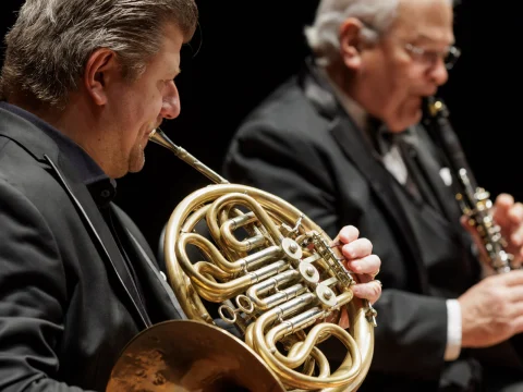 A close-up of two musicians dressed in formal attire playing brass and woodwind instruments during a performance.