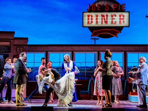 Stage scene set in a diner with actors in colorful costumes; one couple dances in the center as others look on, under a large sign reading "Joe's Pie Diner.