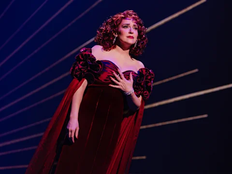 A person in a red velvet gown with puffed sleeves stands on stage against a dark background with diagonal light streaks, holding one hand to their chest.