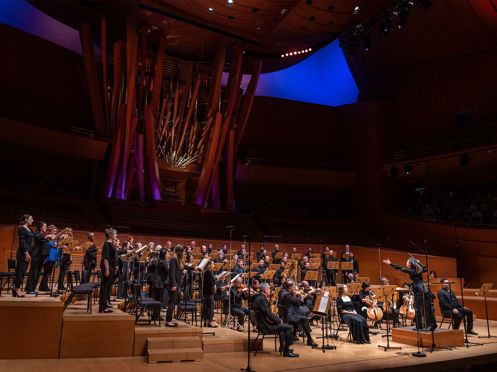 An orchestra and choir perform on stage in a modern concert hall with dramatic lighting and a large organ in the background.
