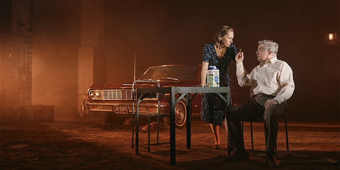 A woman stands by a table facing a seated man who gestures with his hand; a red vintage car is parked behind them in a dimly lit, smoky setting.