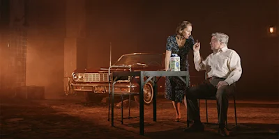 A woman stands by a table facing a seated man who gestures with his hand; a red vintage car is parked behind them in a dimly lit, smoky setting.