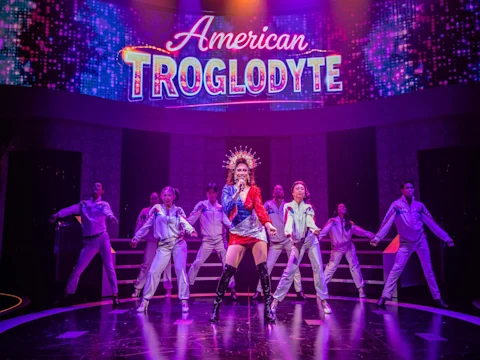 A performer in a sparkling outfit sings at center stage with backup dancers, under a lit sign reading "American Troglodyte" in a vibrant theater setting.