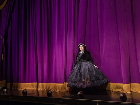 A person in a black Victorian-style dress and bonnet stands alone in front of large, closed purple curtains with gold fringe at the bottom.