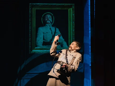 A performer in a tan military-style outfit holds up a telephone; behind them is a large framed portrait of a man in a similar uniform.