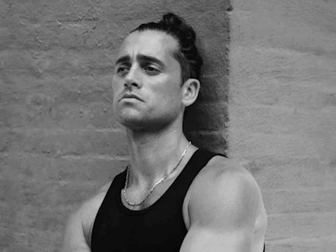 A man with short hair tied in a bun wears a sleeveless black shirt and chain necklace, standing against a textured wall and looking ahead.
