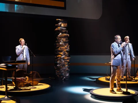 Three men in suits perform on stage with microphones and musical instruments under spotlights.