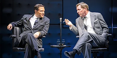 Two men in suits sit across from each other on a stage, engaged in conversation, with a small table and two glasses of water between them.