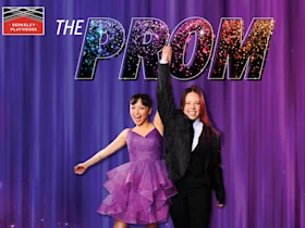 Two people celebrate under the colorful title "The Prom" with one person in a purple dress and the other in a black suit, holding hands and raising one arm. The Berkeley Playhouse logo is in the corner.