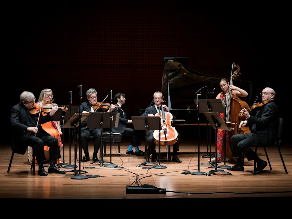 Six musicians perform on stage with string instruments and a grand piano, seated behind music stands and microphones, in a concert hall setting.