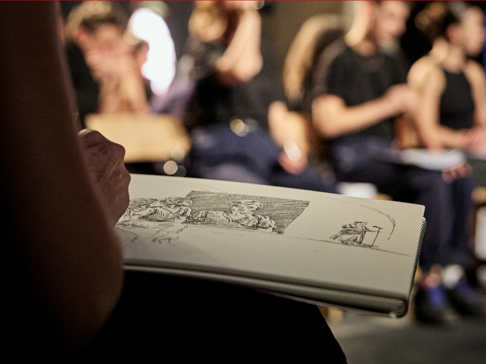 Production photo of Media of Milo Rau & Servane Dècle: The Pelicot Trial in New York, showing A person sketches a group scene in a notebook, with several people sitting and talking in the blurred background.