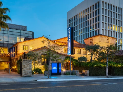 Exterior view of a theater building with illuminated sign reading "Geffen Playhouse" at dusk, featuring stone facade, string lights, and surrounding greenery.