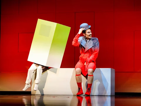 Two performers on stage; one in a large box costume with legs visible, the other in a colorful outfit and bowler hat, sitting on a white block against a red geometric background.