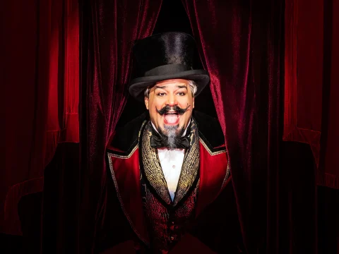 A man dressed in an elaborate circus ringmaster costume stands between red velvet curtains, smiling and wearing a black top hat.