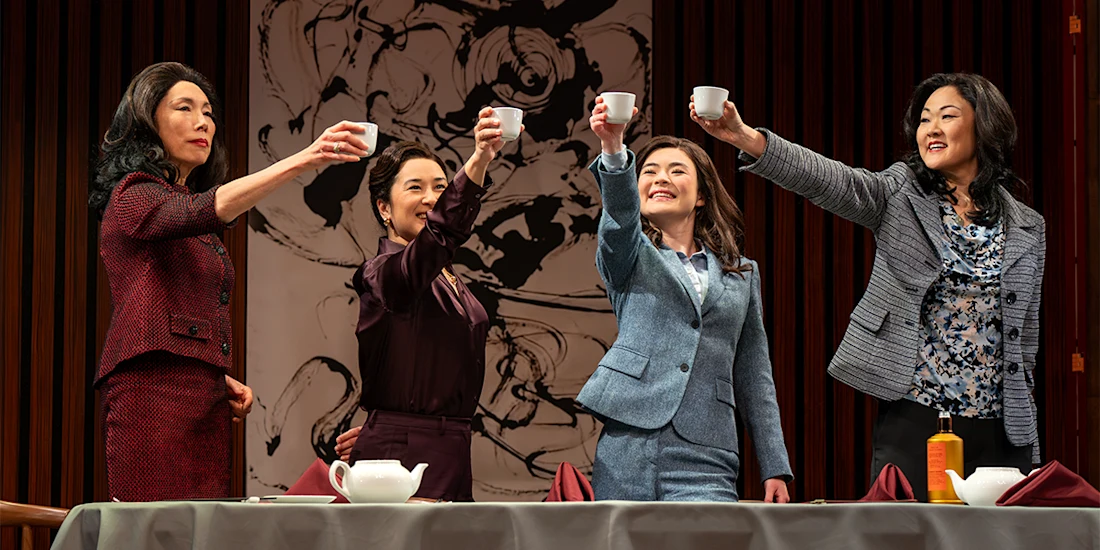 Four women dressed in business attire stand behind a table, raising teacups in a toast, with a large abstract painting in the background.