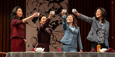Four women dressed in business attire stand behind a table, raising teacups in a toast, with a large abstract painting in the background.