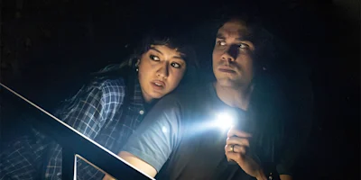 Two people sit in the dark; one holds a flashlight, looking ahead cautiously, while the other appears tense and looks over their shoulder.
