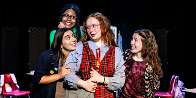 Four people stand closely together indoors, smiling and posing. The person in the center wears glasses and plaid, while the others appear to interact with them. Red chairs are visible in the background.