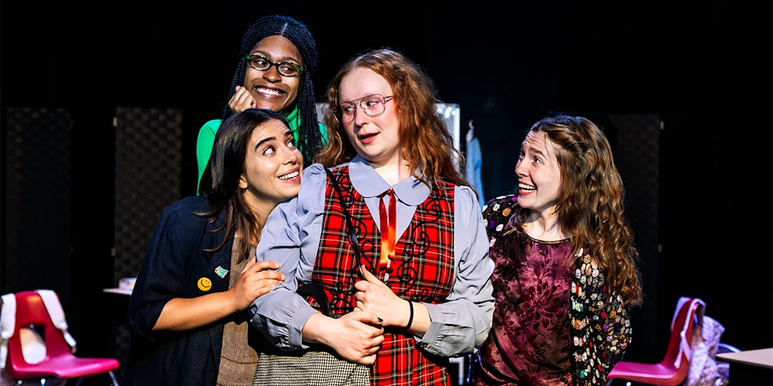 Four people stand closely together indoors, smiling and posing. The person in the center wears glasses and plaid, while the others appear to interact with them. Red chairs are visible in the background.