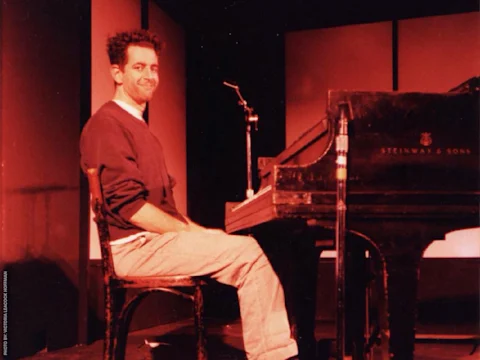 A person sitting at a Steinway & Sons piano on a stage, wearing a casual outfit. The lighting is red.