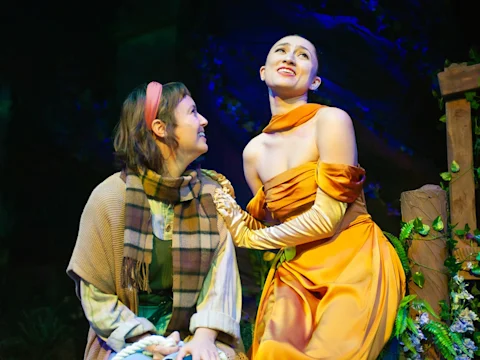 Two actors on stage: one in an orange gown and gloves sings or speaks passionately, while the other, wearing a scarf and shawl, looks at her attentively.