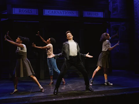 Four actors perform on stage in front of a flower shop set, with one person in the center posing dramatically and three others striking expressive poses around them.