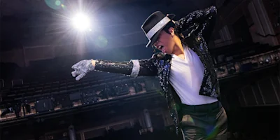 Person wearing a sequined jacket, white glove, and black hat poses on stage under a spotlight, evoking a classic Michael Jackson dance move.