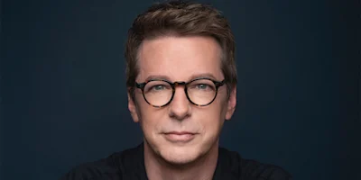 A man with short brown hair and glasses looks directly at the camera against a dark background.
