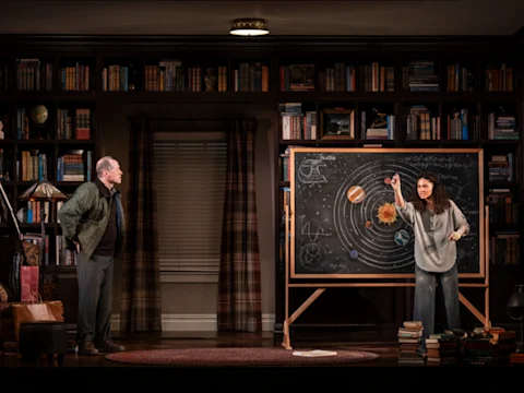 Two people stand in a library; one gestures at a large chalkboard displaying a solar system diagram, while the other looks on.