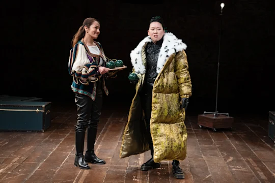 Two people on stage: one holds a tray with green balls, wearing a striped top and leather pants; the other in a gold coat with fur trim, holding a ball, stands confidently.