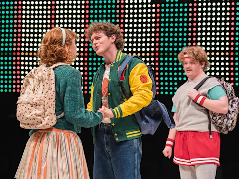Three young people wearing 1980s-style costumes stand on stage; two hold hands and face each other, while a third looks on, with a backdrop of colorful lights behind them.